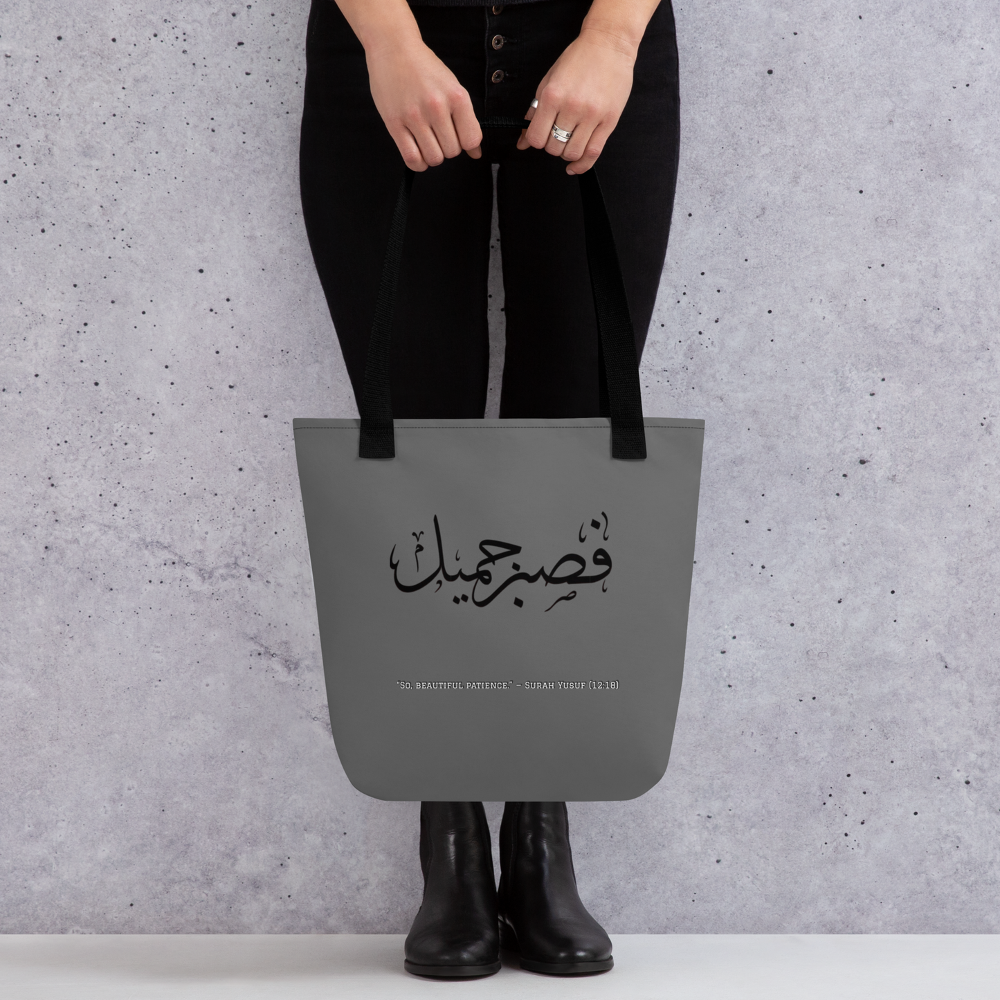 Person holding a gray tote bag with Arabic text against a gray background